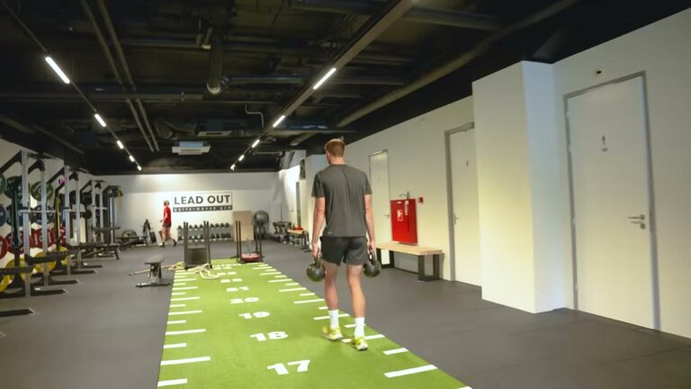 Atleet loopt een farmers carry met twee kettlebells over de groene HYROX-trainingsbaan
