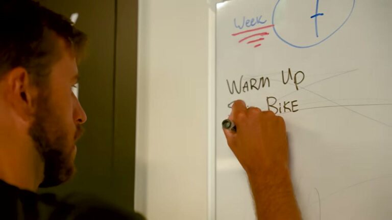 Coach schrijft een HYROX-weekschema op een whiteboard met warming-up en bike